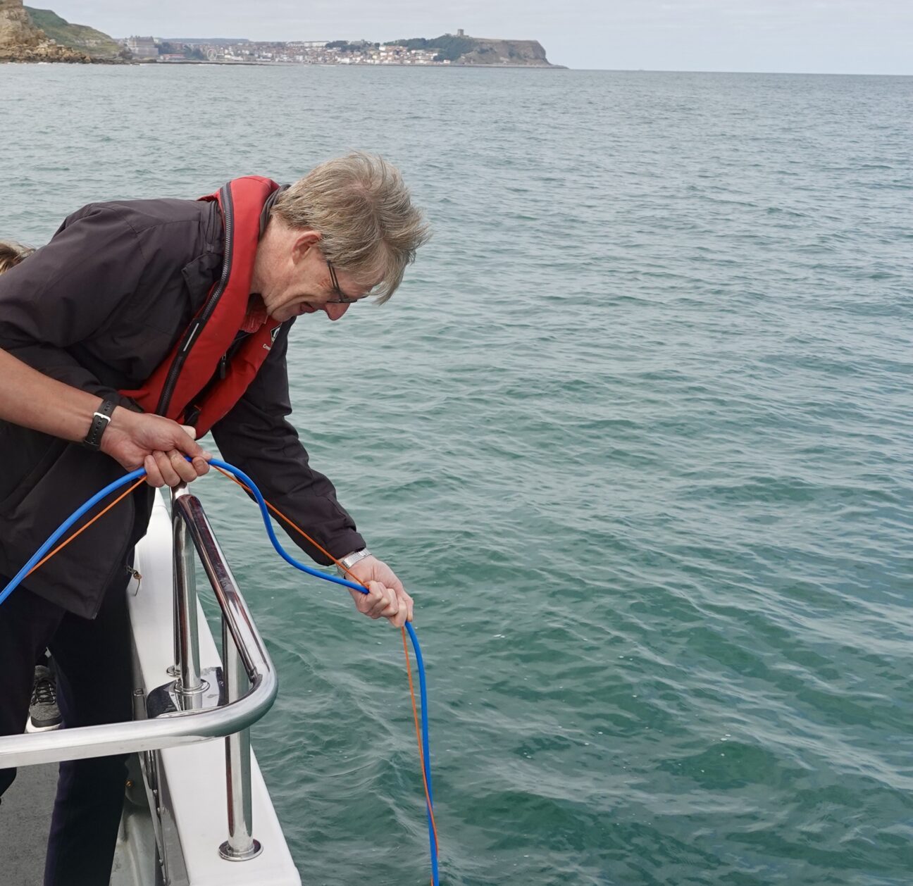 A man wearing a buoyancy aid lowers blue rope over the side of the boat into the sea.