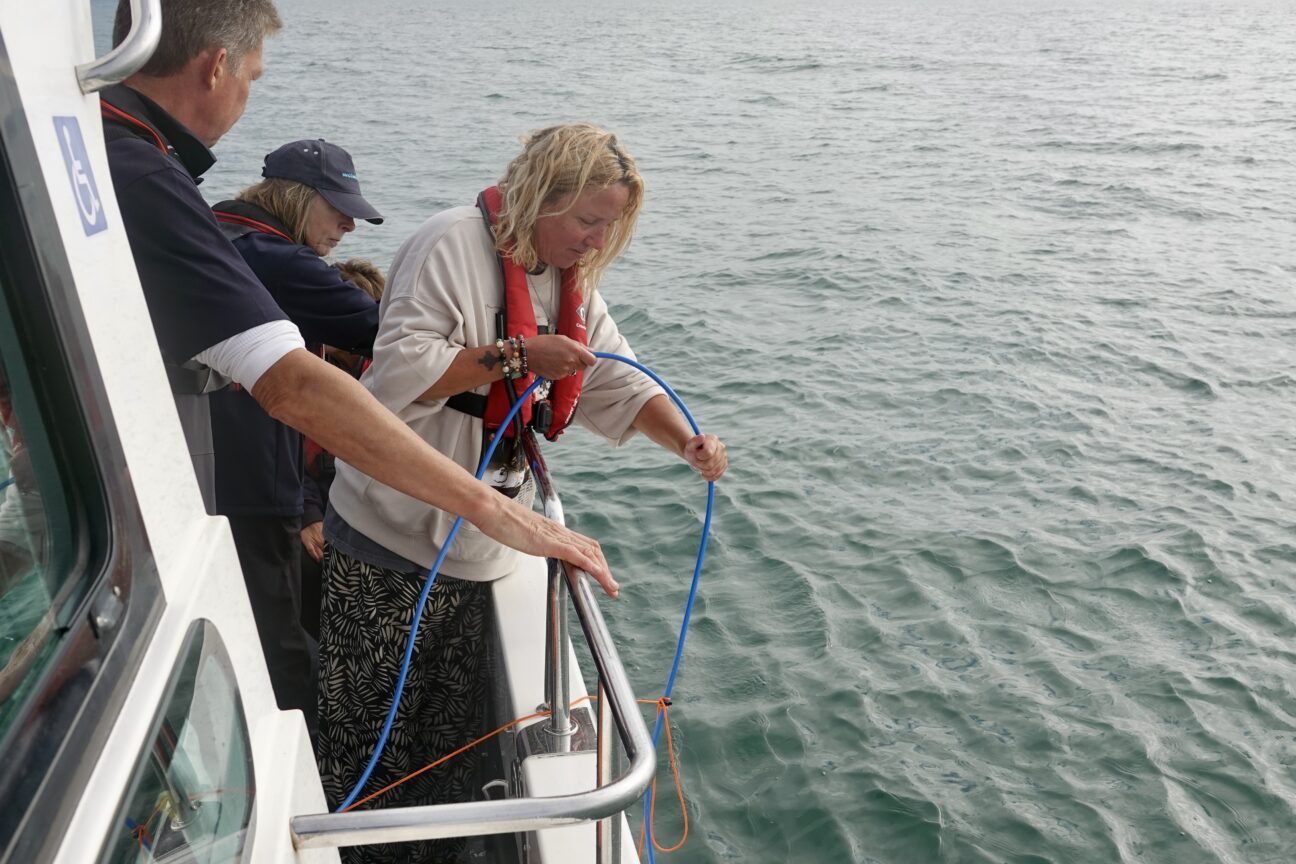 A woman wearing a buoyancy aid lowers blue rope over the side of the boat into the sea.