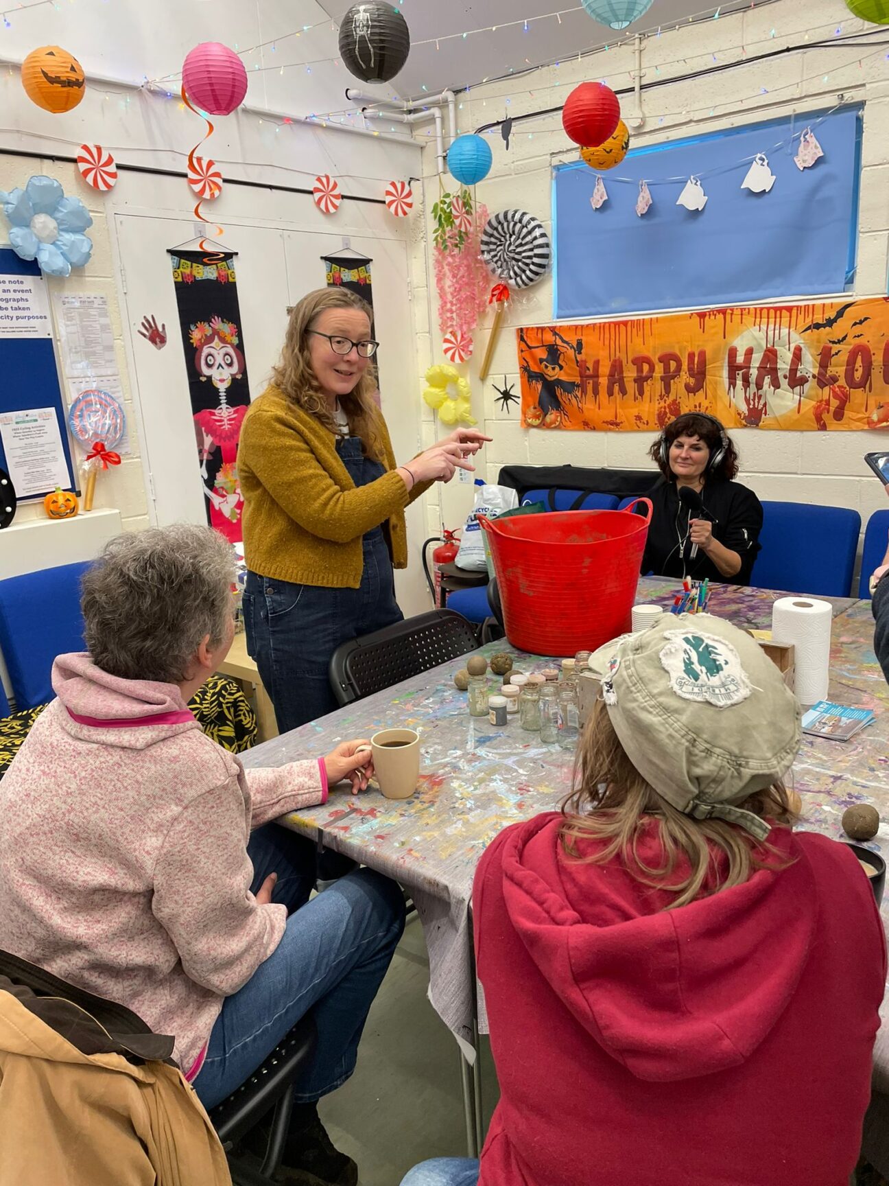 Woman wearing glasses and yellow cardigan stands in front of a table surrounded by a group of people. She is explaining how to make a Dorodango ball.