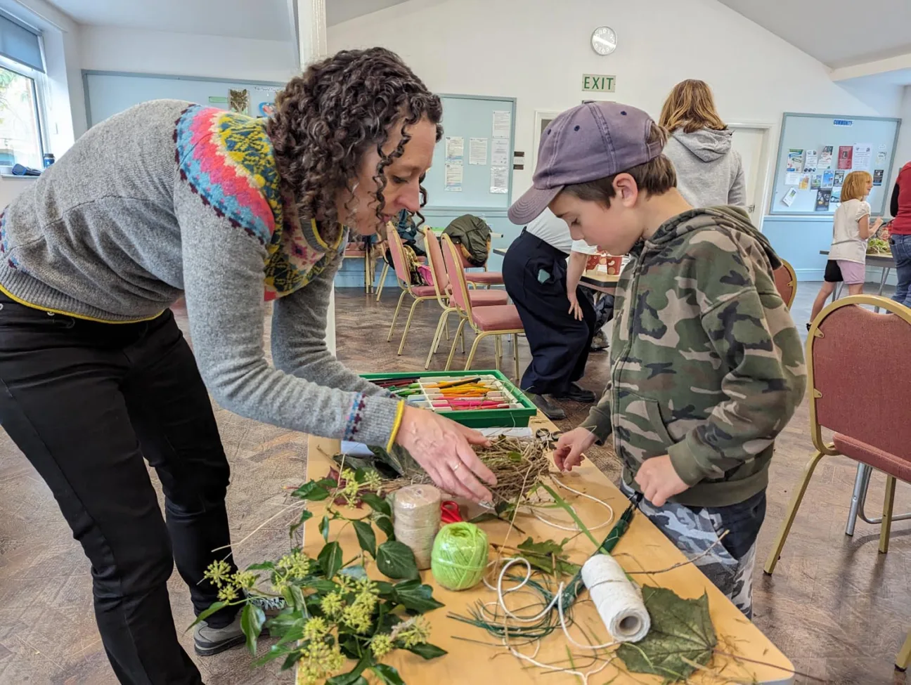 Woman leans down to a table to help a child create a circular habitat for wildlife using sticks.