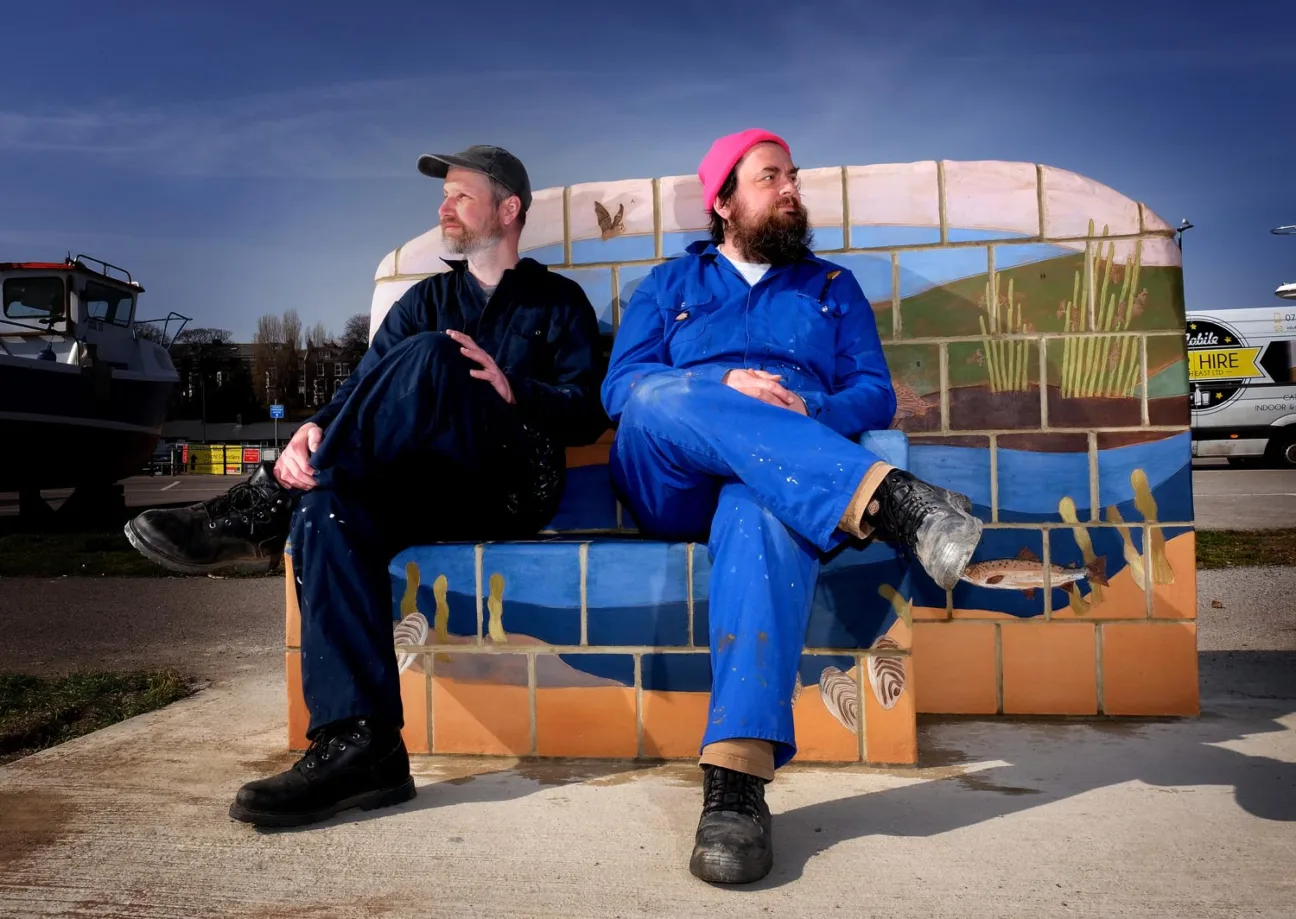 The artists Juneau Projects, both wearing blue work overalls, sitting on one of their sculptural benches, decorated with imagery of animals and the natural landscape.