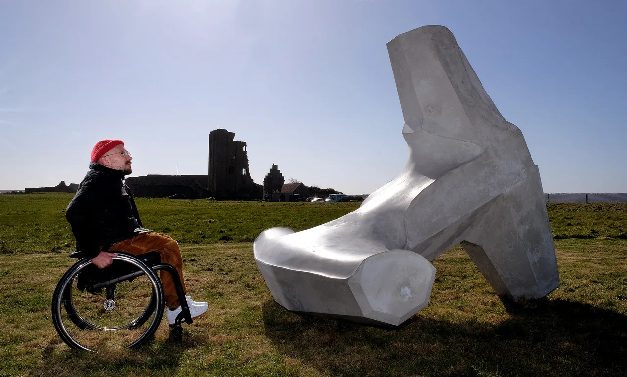 The artist Ryan Gander sitting in a wheelchair next to his sculpture with Scarborough castle in the background