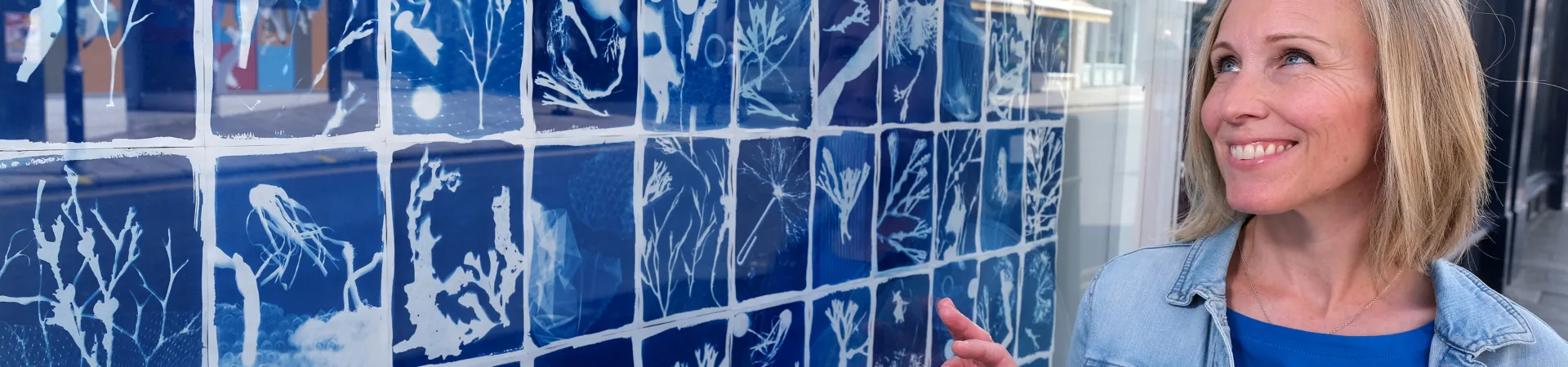 Artist Jacqui Barrowcliffe in front of community made cyanotypes displayed in an empty shop window. The rectangular cyanotypes have a blue background with a white outline of the printed item.