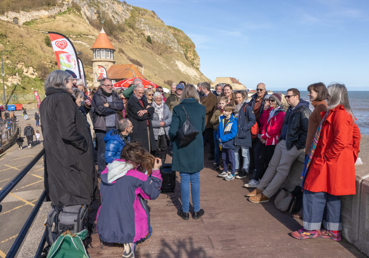 Professor Juliet Brodie in front of a crowd on Scarborough Harbour, talking about the role of seaweed in the environment. The crowd are faced towards her, listening.