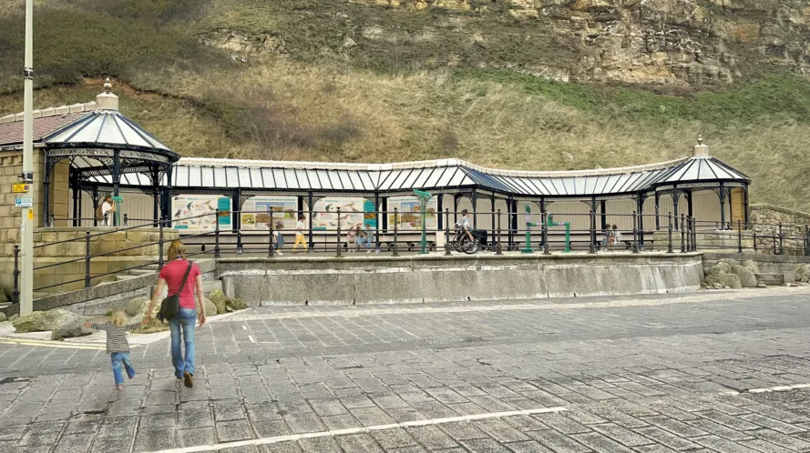 An imagined image of what the sea watching station will look like from the road. The shelter has a roof, there are black railings in front of the green telescopes. There is an adult and a child crossing the road holding hands in front of the shelter.