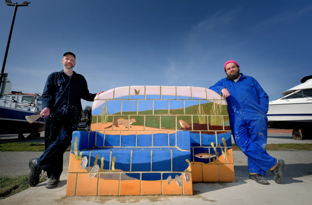 Two members of Juneau Projects stand with one of their colourful benches, depicting estuary nature, including freshwater pearl mussels and fish.