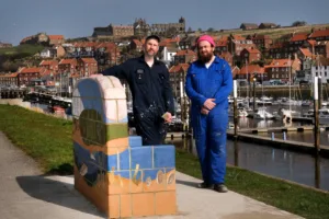 The artists Juneau Projects, both wearing blue work overalls, stand on either side of one of their sculptural benches, decorated with imagery of animals and the natural landscape. Whitby Abbey stands above the town in the background.