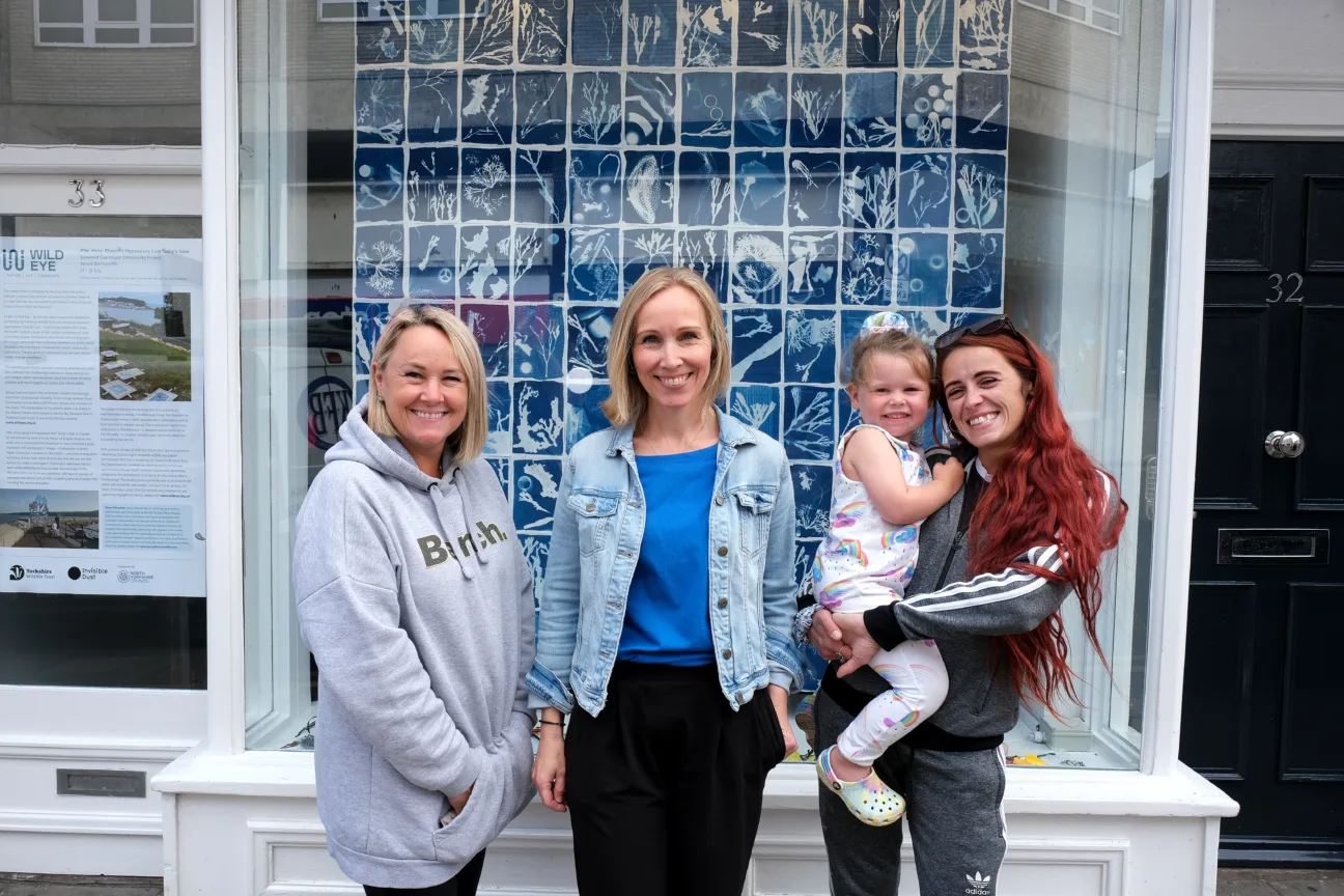 Artist Jacqui Barrowcliffe stands between two women, one holding a small child. All smile into the camera. They stand in front of a white-framed shop window with a display of blue and white sun prints.
