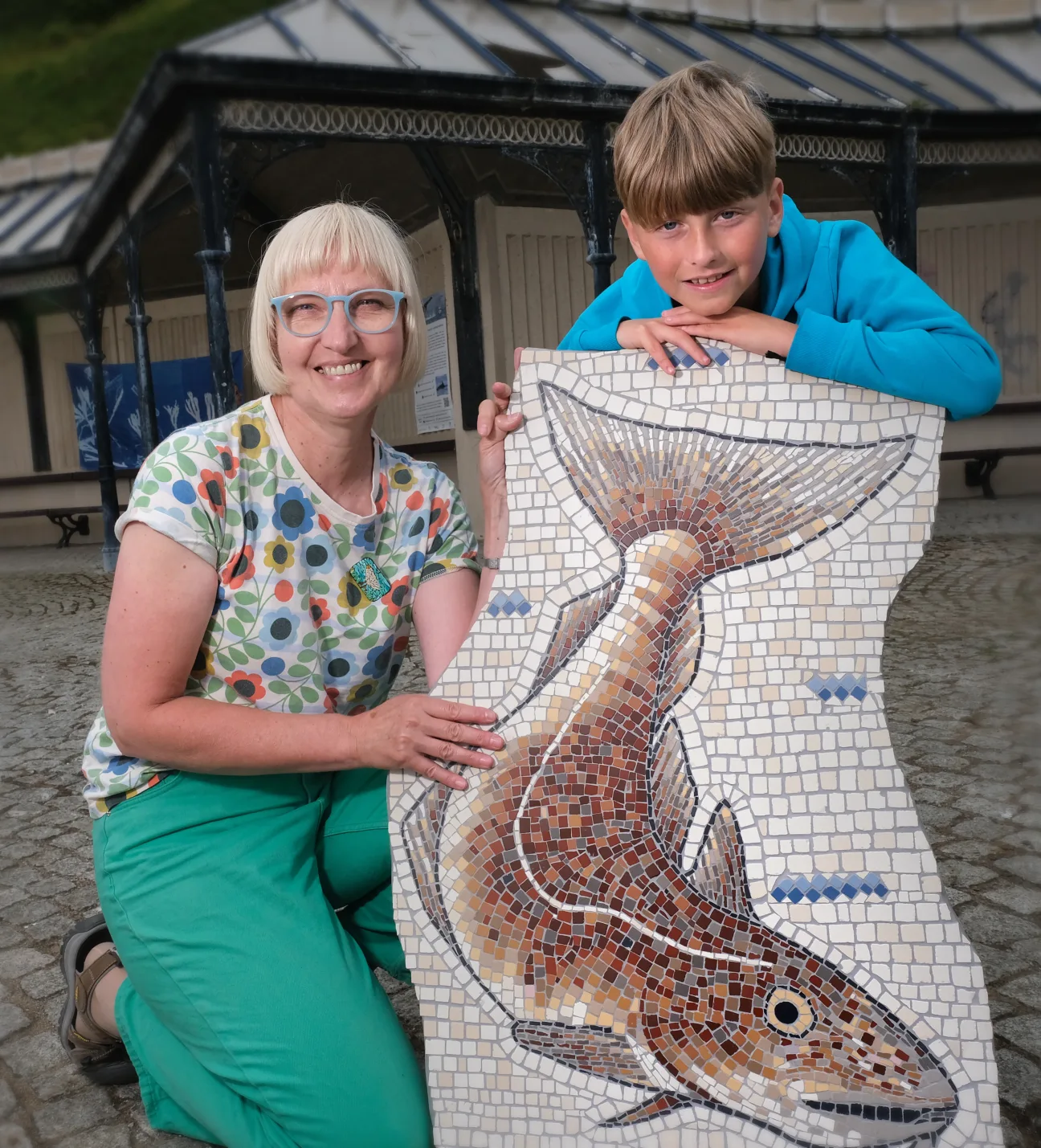 Mosaic artist Coralie Turpin kneels next to a young participant. They smile into the camera whilst presenting a sample of mosaic depicting a fish.